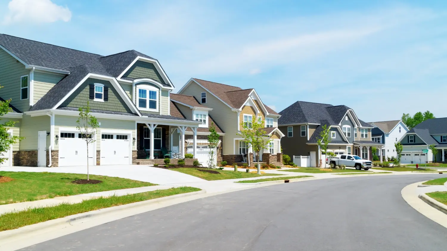 A curving street of new two-storey suburban houses with attached garages and young trees, under a clear sky.